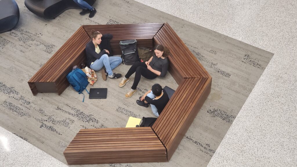 One of the first SpEED meetings. Christina, Sofia, and Rachel sit on the carpet inside of an open wooden hexagon bench, deep in conversation. The photo looks down on this scene from a floor above.