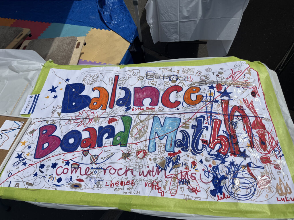 Photograph of the Balance Board Math table at science fair. A large piece of white paper covers the table. "Balance Board Math" is written in blue, pink, orange, green, and red bubble letters, with "come rock with us" written underneath in red. Children have added doodles and tags all around the letters in red, blue, and gold including swirls, phrases like "is the best" and "great job guys", signed names, swirls, and cartoon humans and animals. 