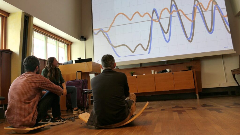 Two men sit on different types of balance boards: a large curved piece of wood, and one with a flat surface and rounded bottom. In front of them looms a giant display showing variable orange, blue, and black graph lines that become more synchronized towards the end of the graphing area.