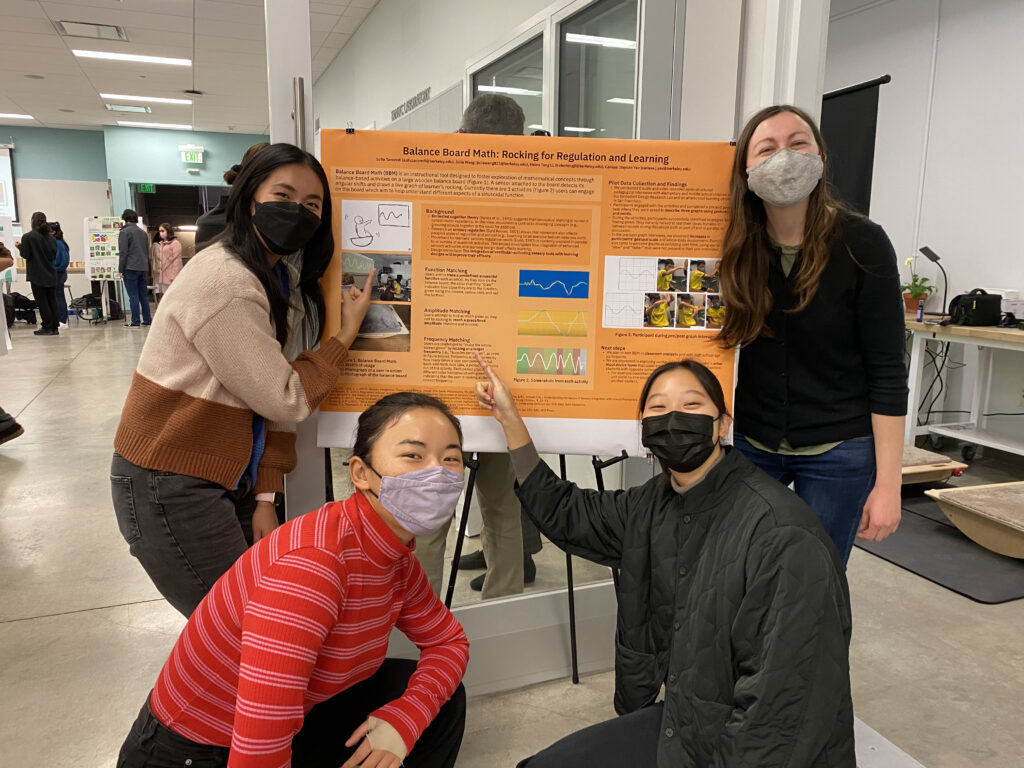 Four masked women surround an orange showcase poster titled "Balance Board Math: Rocking for Regulation and Learning." Helen and Sofia stand on either side and Carissa and Julia squat in front. The poster shows blurbs about three different activities each with a colorful graph and images of a participant gesturing. Julia points to the third activity. Two balance boards can be seen behind Sofia in the background. Behind Helen, other showcase posters are visible in the background. 